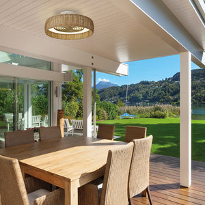 A wicker ceiling fan in a covered, external terrace area above alarge wooden table with six wicker chairs around it. The colours are neutral and in the background is a lawn and a lake.